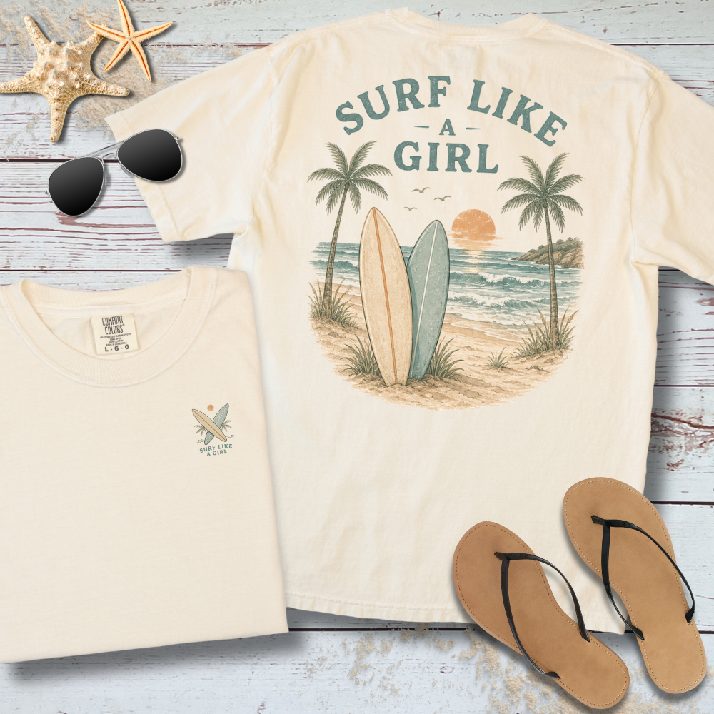 Beige t-shirt with 'Surf Like a Girl' design, sunglasses, and flip-flops on a wooden surface.