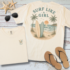 Beige t-shirt with 'Surf Like a Girl' design, sunglasses, and flip-flops on a wooden surface.