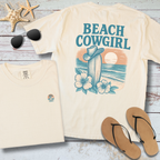 Beach-themed t-shirt with 'Beach Cowgirl' design on a wooden surface with sunglasses and flip-flops.
